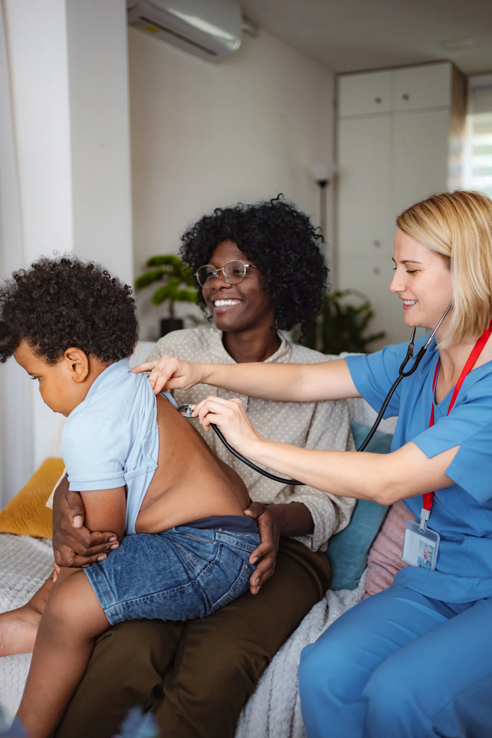 Nurse examining boy with stethoscope while mother holds son on sofa